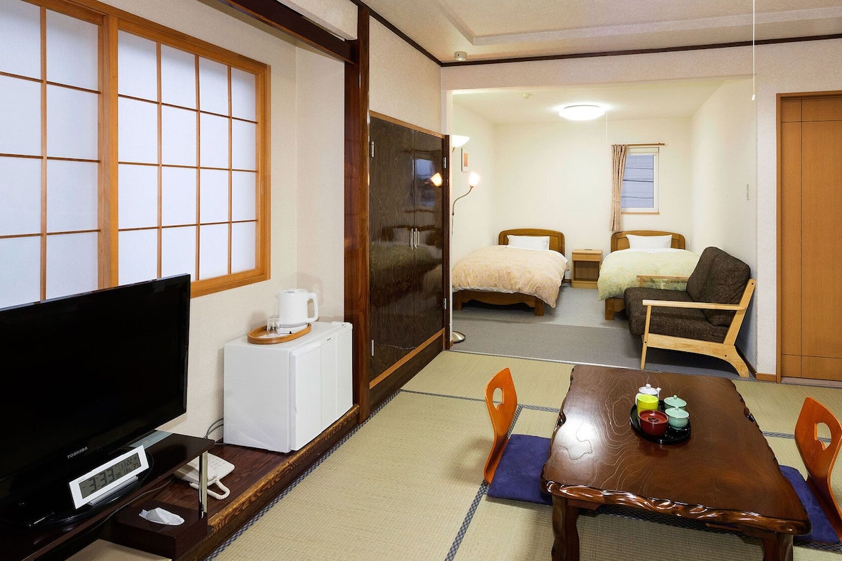 Ett traditionellt japanskt rum med tatamimattor på golvet, en tv, ett träbord med ett teservis och två sängar.