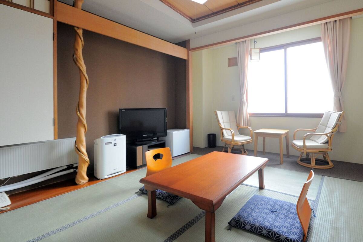 Ein Zimmer mit Tatami-Böden, einem Fernseher, einem kleinen Tisch, zwei Stühlen und einem Futon.
