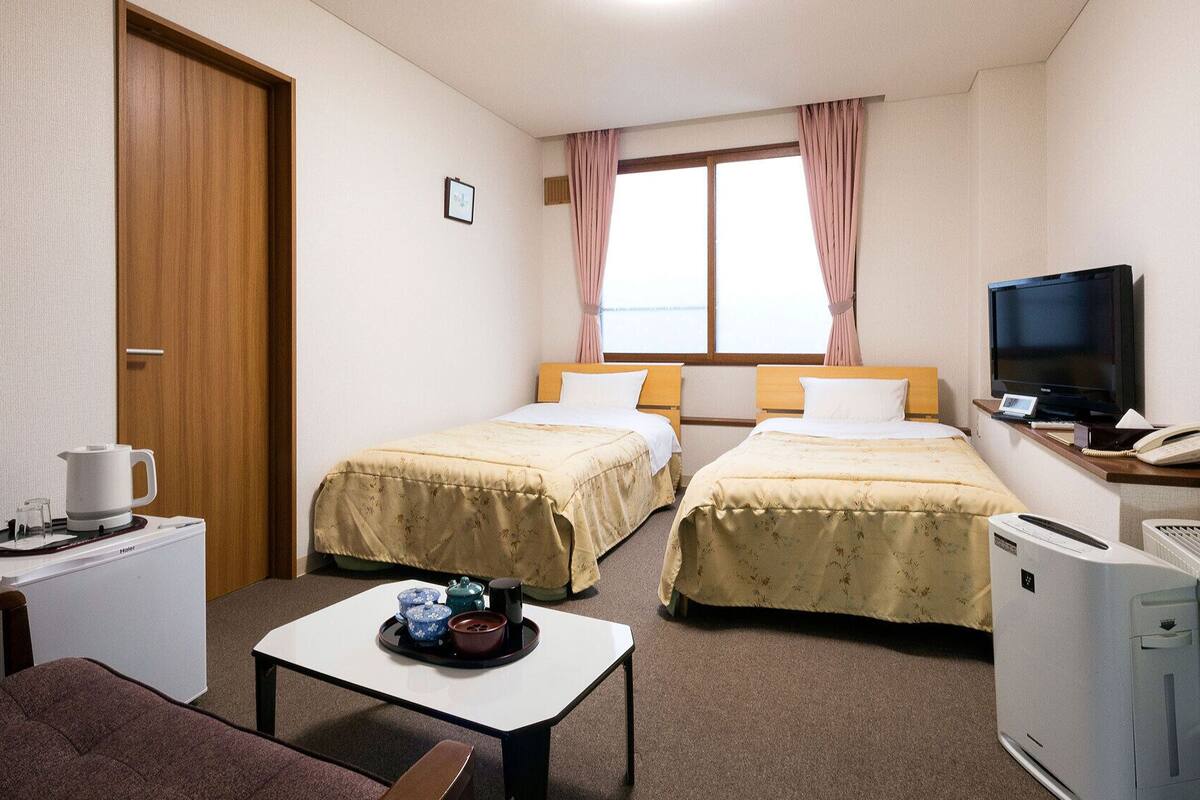 Ein Hotelzimmer mit zwei Betten, einem Fernseher, einem Schreibtisch mit Wasserkocher, einem kleinen Tisch mit Geschirr und einer Heizung.