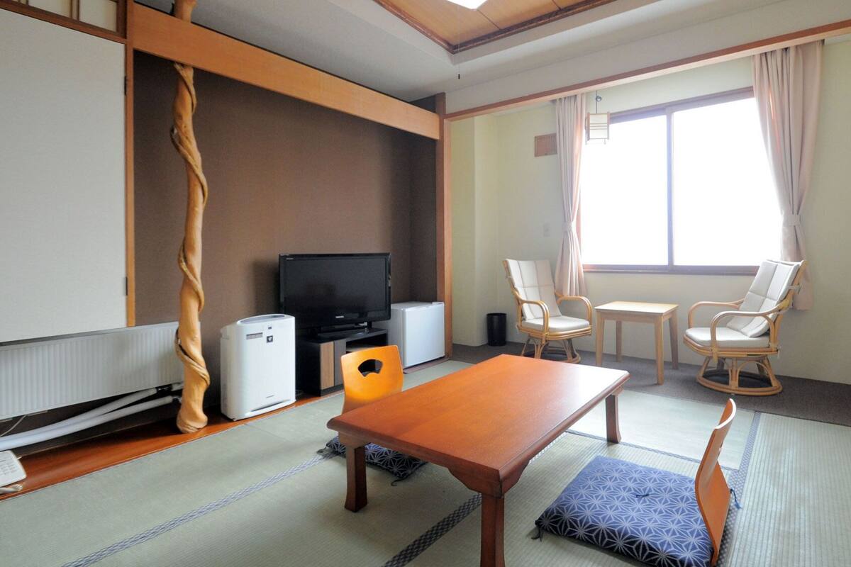 Ein Zimmer mit Tatami-Boden, einem Fernseher, einem kleinen Kühlschrank, einem Holztisch, zwei Stühlen und einem fenster mit Vorhängen.