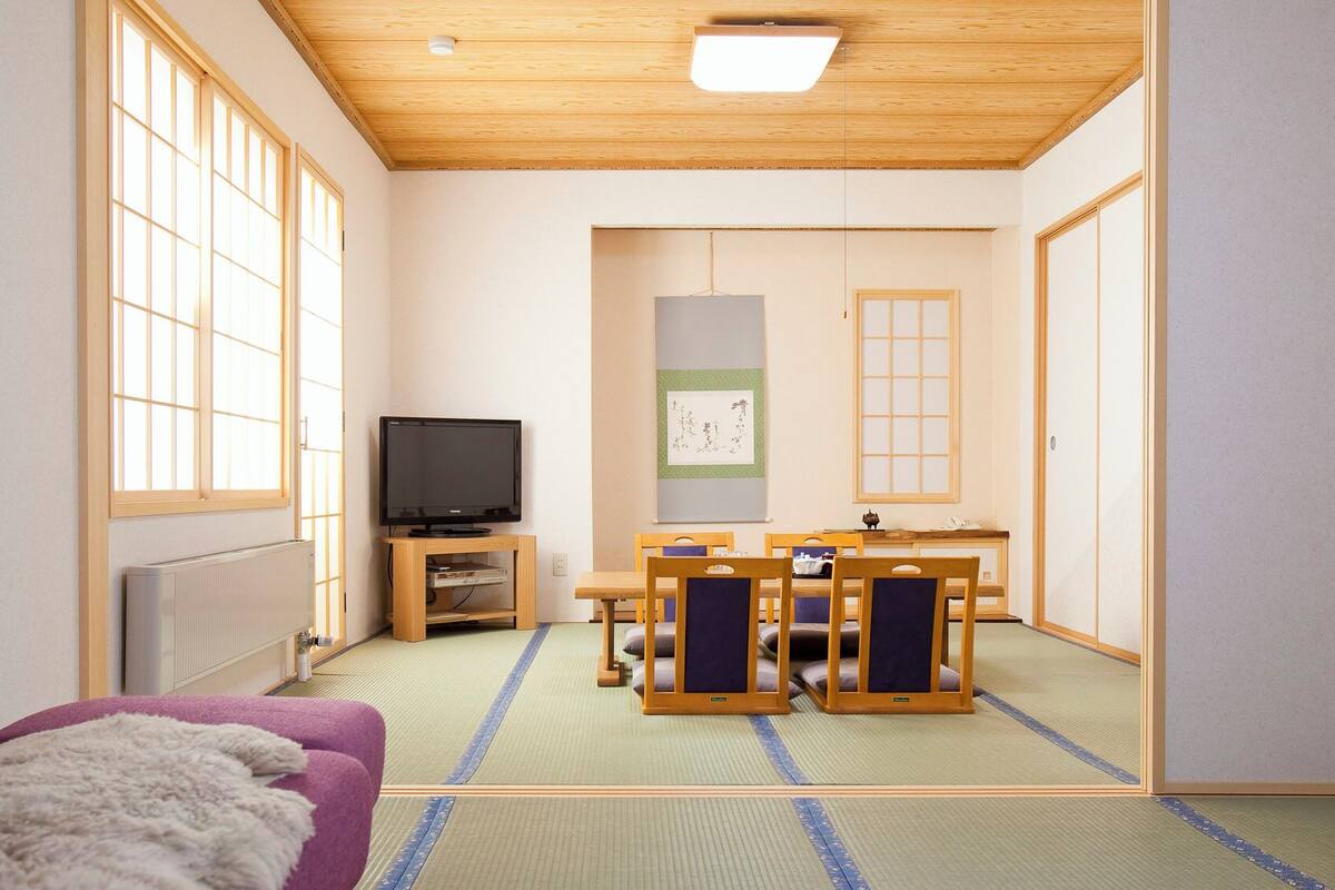 Ein traditioneller japanischer Raum mit Tatami-Boden, einem Fernseher, einem Tatami-Tisch mit Stühlen und einem Shoji-Bildschirm.