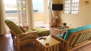 Ein Zimmer mit einer Holzsofa, einem Holzsesseln, einem kleinen Tisch, einem Fernseher auf einem Standfuß und einem Fenster mit Blick nach draußen.