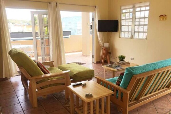 A room with a wooden sofa, a wooden armchair, a small table, a television on a stand, and a window with a view of the outside.