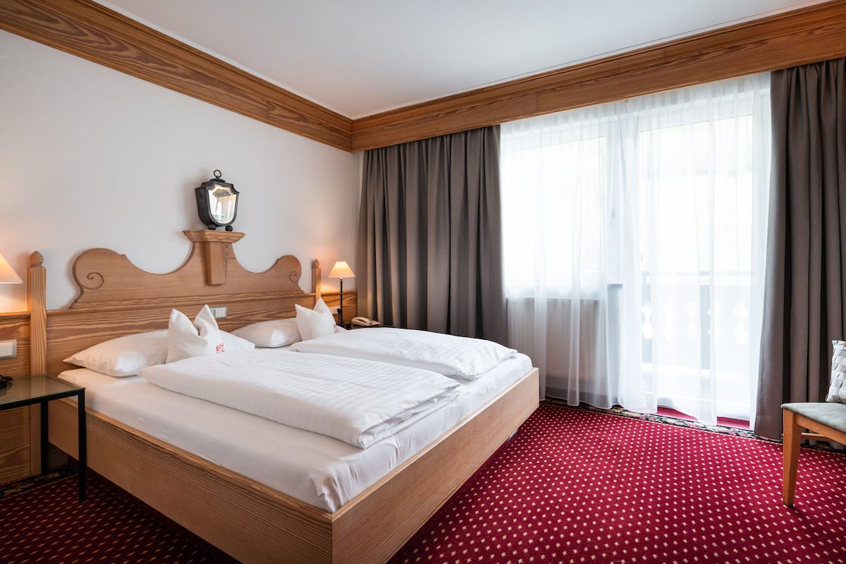 Habitación de hotel con una cama grande, cabecera de madera, dos lámparas de noche, una mesita de noche, piso cubierto de alfombra roja y una ventana con cortinas.