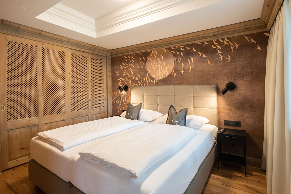 Cama doble con cabecera de madera, mesita de noche con lámpara, y pared decorada con un mural de pájaros.