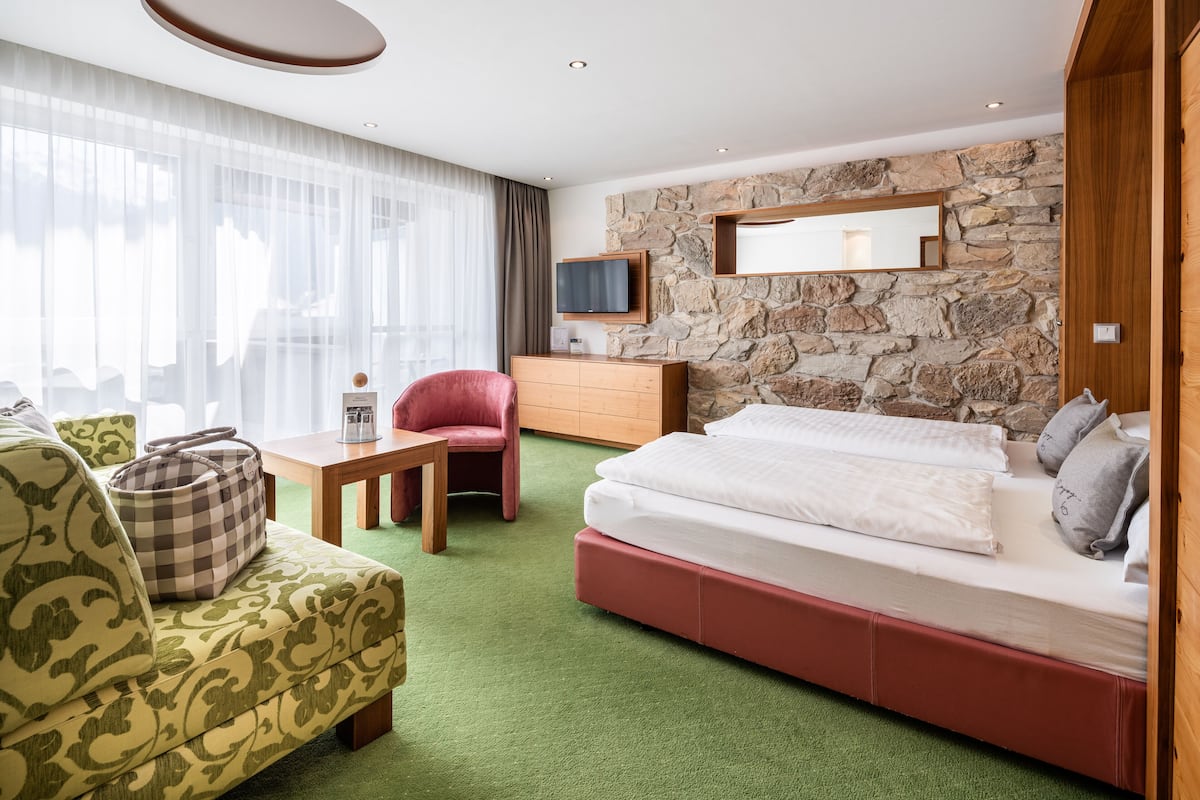 Habitación de hotel con alfombra verde, cama con cabecera roja, cómoda de madera, una silla roja y un ventanal con cortinas translúcidas.