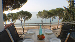 Une table dressée pour quatre personnes, avec des assiettes, des verres et une bouteille, donnant sur une plage bordée d’arbres et sur la mer.