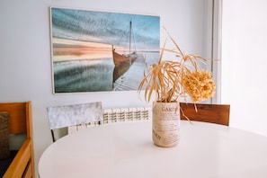 A dining area with a white table, a vase with dried plants, a wooden chair, and a painting of a boat on a pier. - Agi Marina 6 Apartamento for rent in Roses Girona. (Roses)