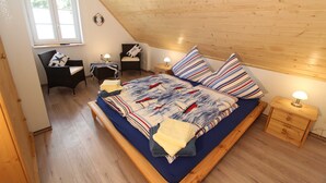 Ein Schlafzimmer mit schrägen Wänden, einem Bett mit gestreifter Bettwäsche, zwei Nachttischen mit Lampen, einem Sessel und einem kleinen Schreibtisch mit Stuhl.
