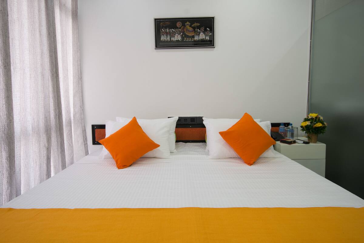 A hotel room with a bed, white bedding, orange pillows, a nightstand with a vase of flowers, and a framed picture on the wall.