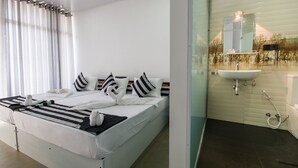 Una habitación de hotel moderna con una cama bien hecha, un ventanal con cortinas, un baño con lavabo y espejo, y piso de baldosas.