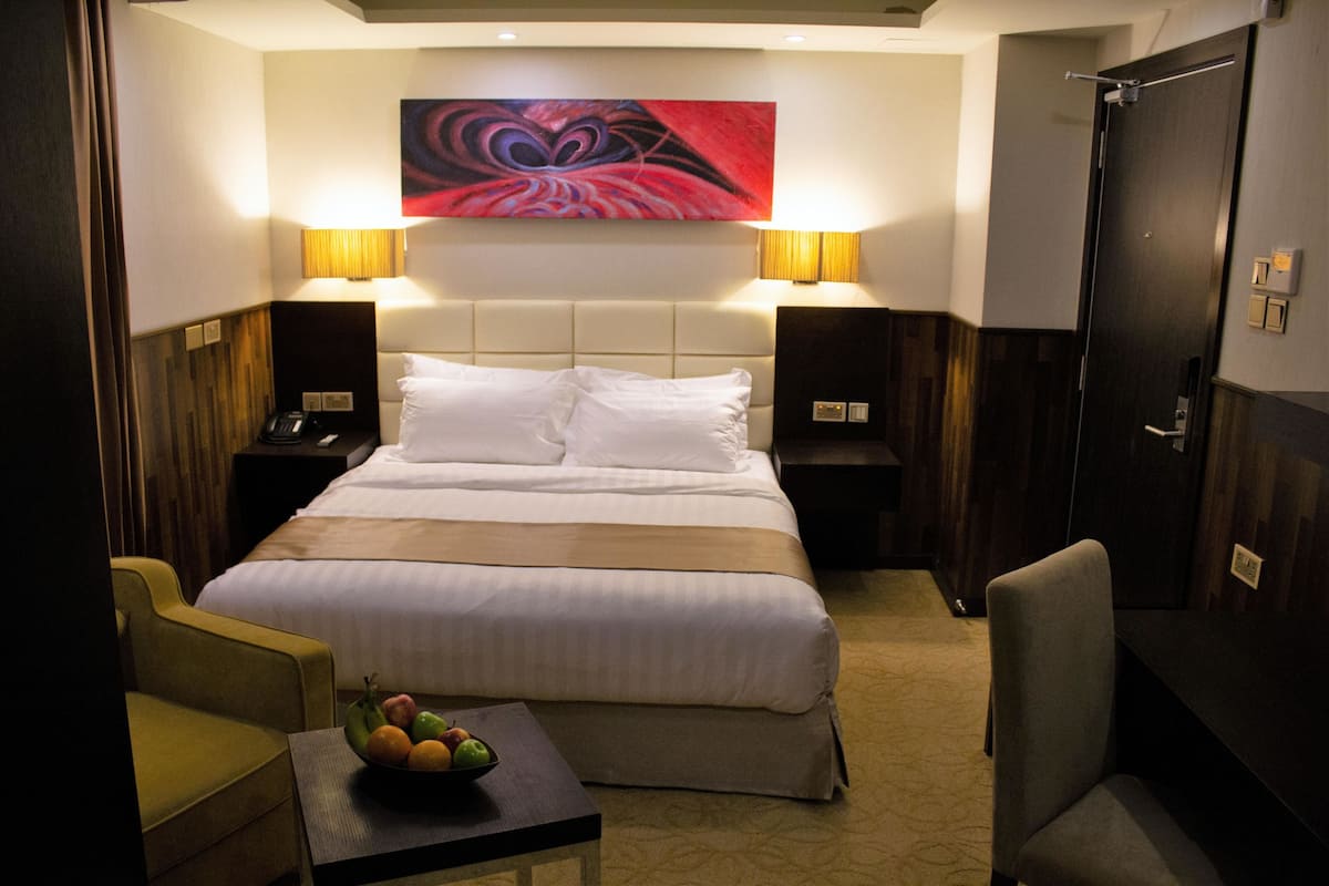 A hotel room with a bed, two bedside lamps, a fruit bowl on a side table, and a painting above the bed.
