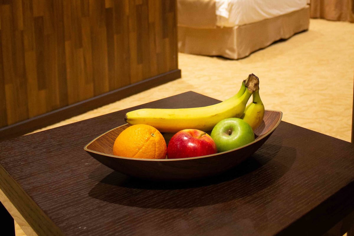 A hotel room with a bed, a wooden headboard, a bedside table with a fruit bowl, and a view of the room's interior.