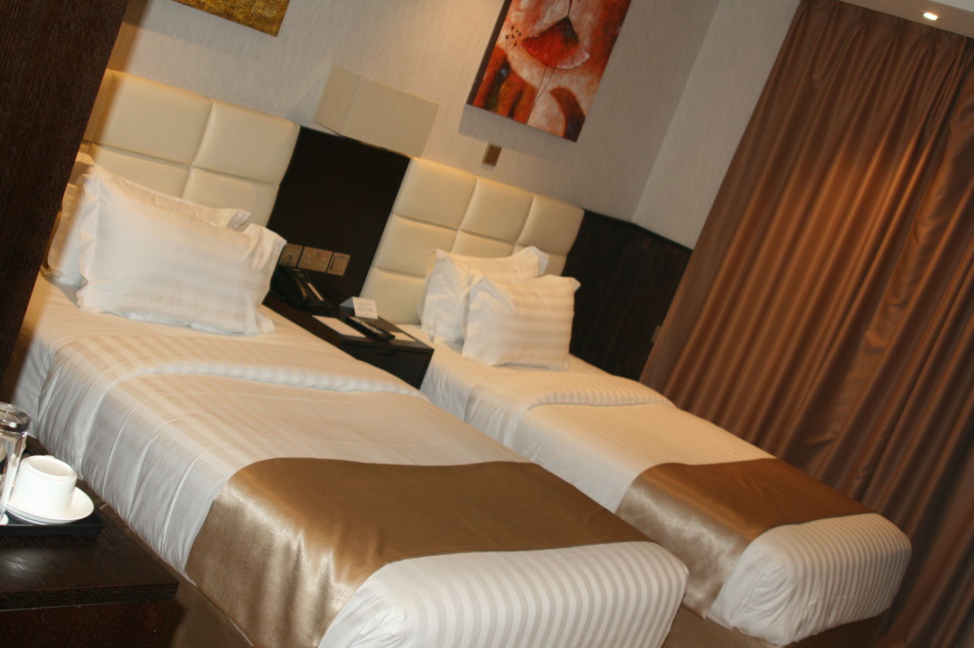 A hotel room with two beds, a bedside table with a cup and saucer, and a painting on the wall.