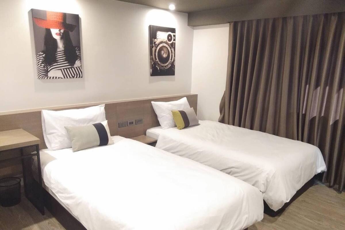 A hotel room with two single beds, a wooden headboard, a small table, and a trash bin.