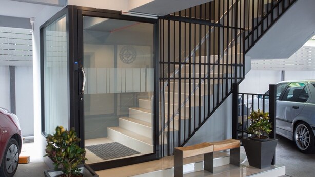 A modern interior with a glass door entrance, a staircase with metal railings, and a bench. - Toptel Thaphra (Bangkok)