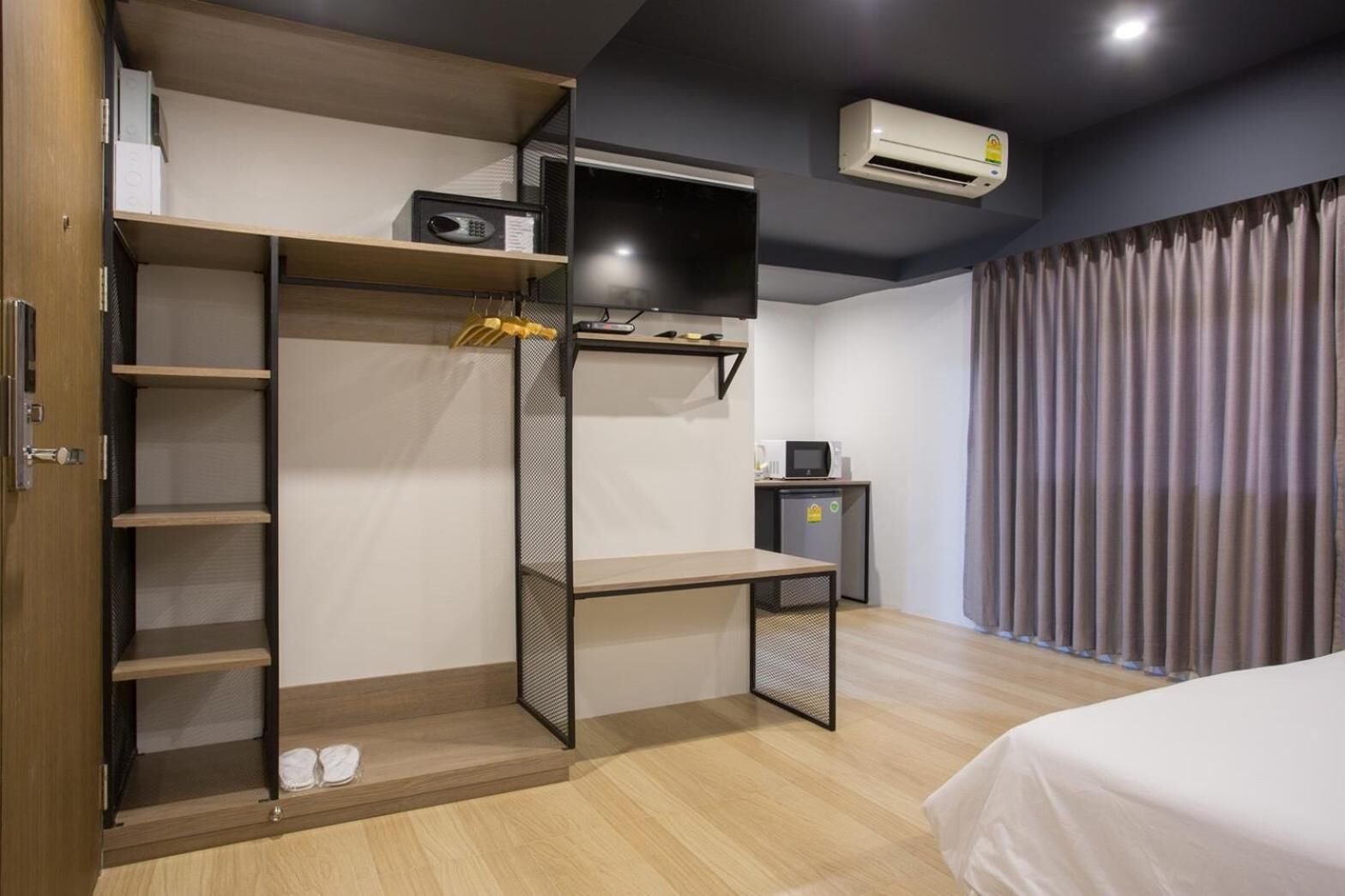 A bedroom with a bed, a wardrobe, a desk, a small kitchenette, and an air conditioning unit.