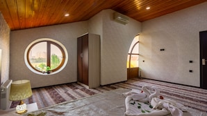 Une chambre d’hôtel avec un plafond en bois, une fenêtre ronde, un lit recouvert d’une couvre-lit à motifs et une table de chevet avec une lampe.