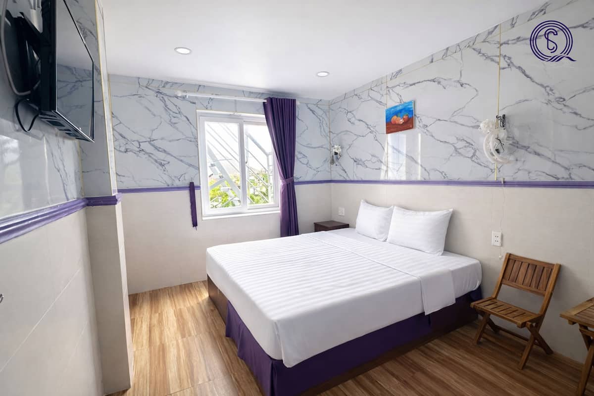 A hotel room with a bed, wooden floor, a TV, a window with curtains, and a marble wall.
