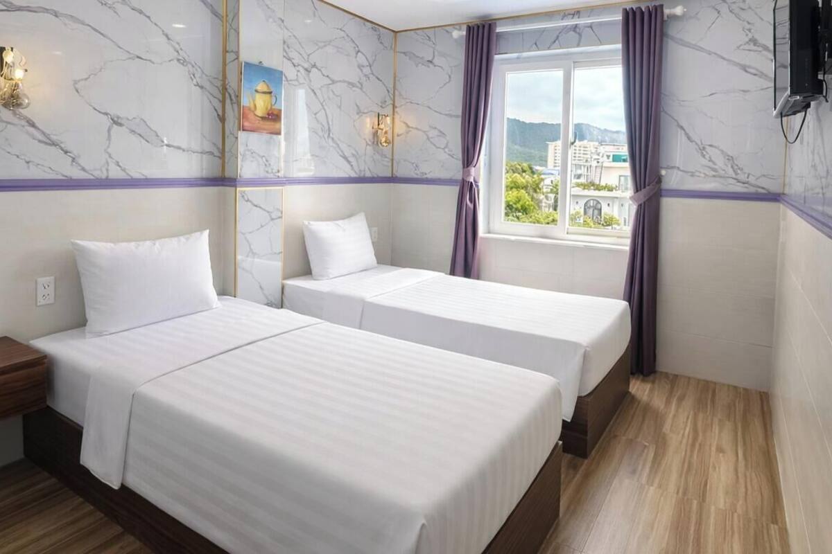 A hotel room with two beds, a nightstand, a window with curtains, marble walls, and a view of buildings.