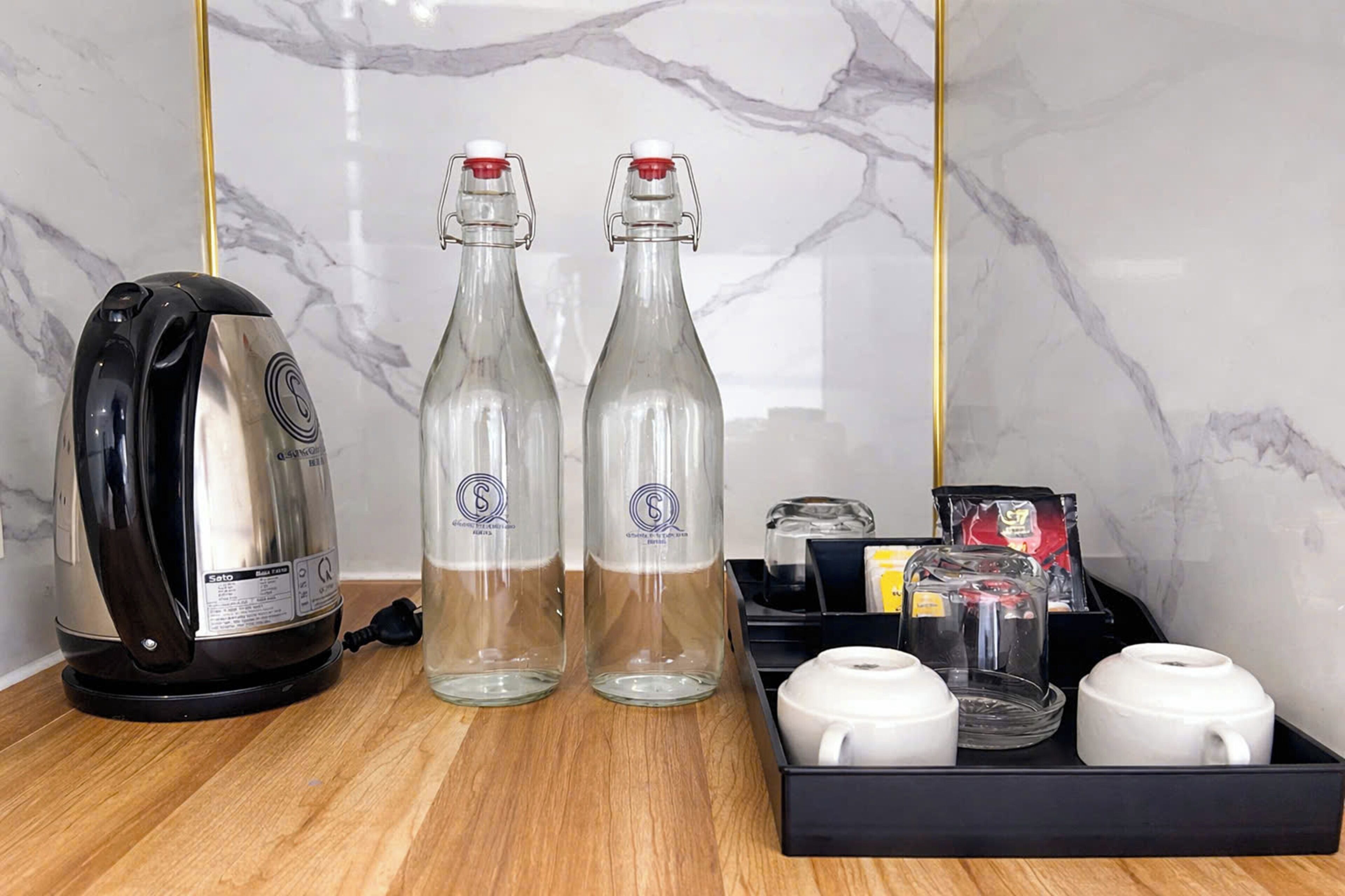 Un área de cocina con un hervidor, dos botellas de vidrio, un juego de té y una bandeja negra con una bolsita de té y una caja.