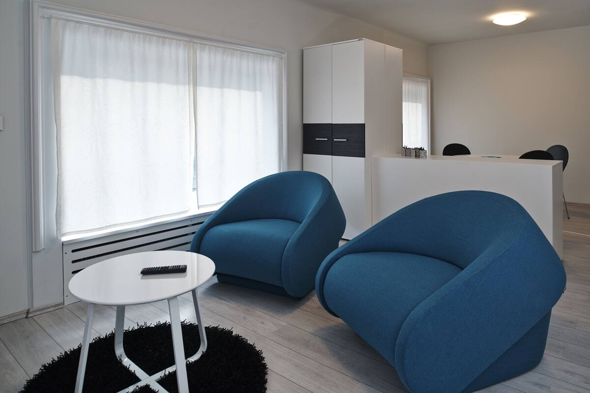 A modern room with two blue armchairs, a small white table, and a kitchenette in the background.