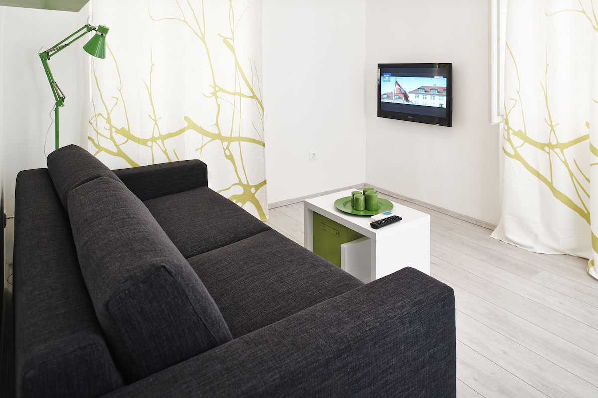 A modern living room with a grey sofa, a green floor lamp, a small white side table with a remote control, and a TV on the wall.