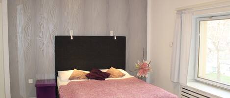 Un dormitorio moderno con una cama grande, una mesita de noche, un jarrón decorativo y vistas a través de una ventana.