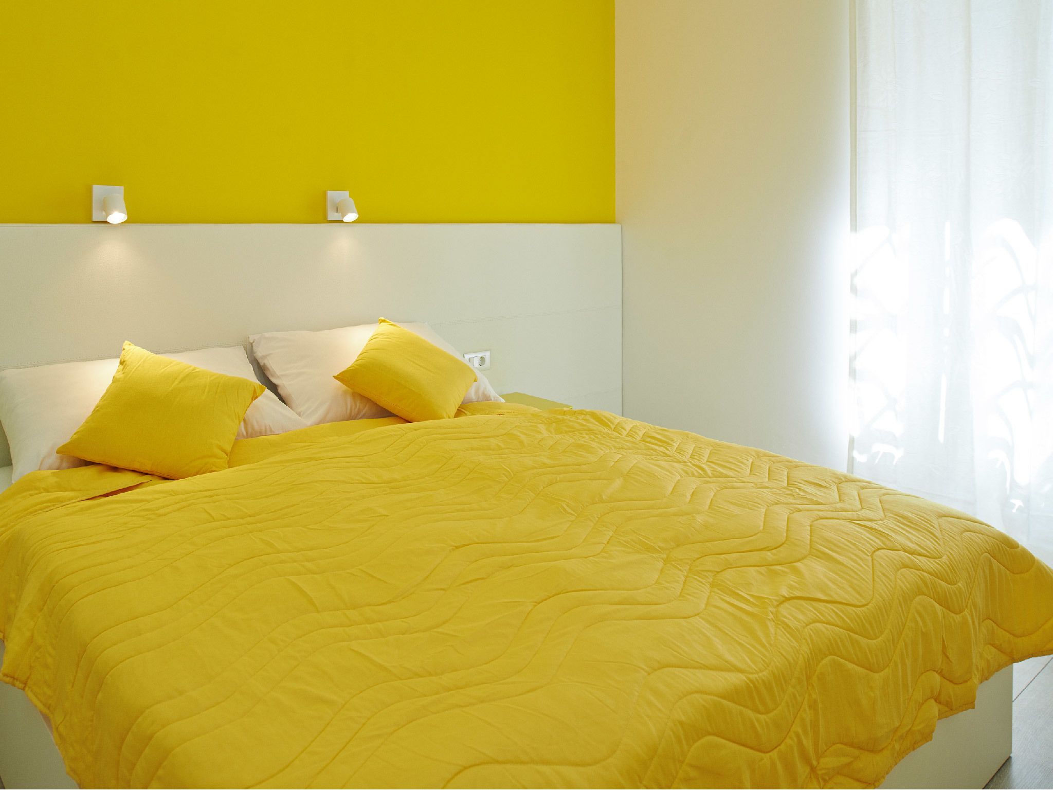 Un lit avec une couette et des oreillers jaunes, une tête de lit blanche et un mur d’accent jaune.