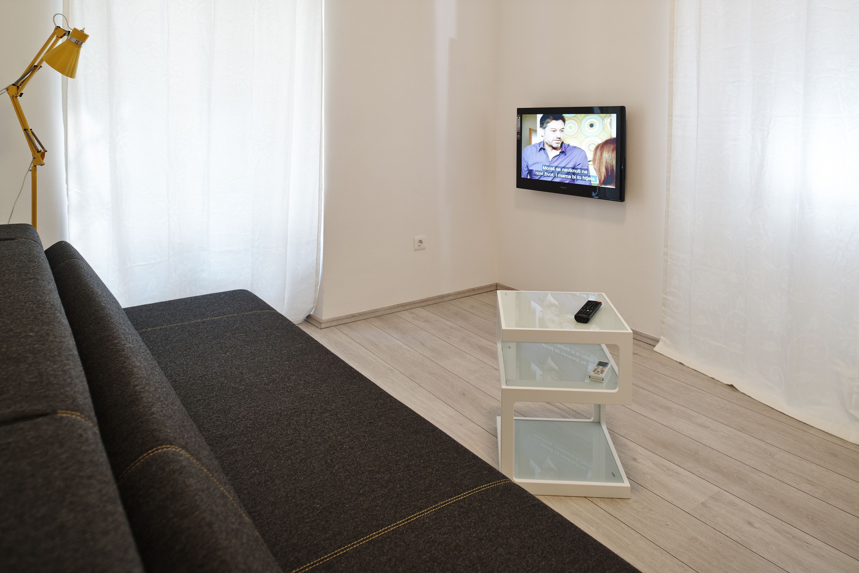 A living room with a grey sofa, a white TV on the wall, and a small white side table with a remote control.