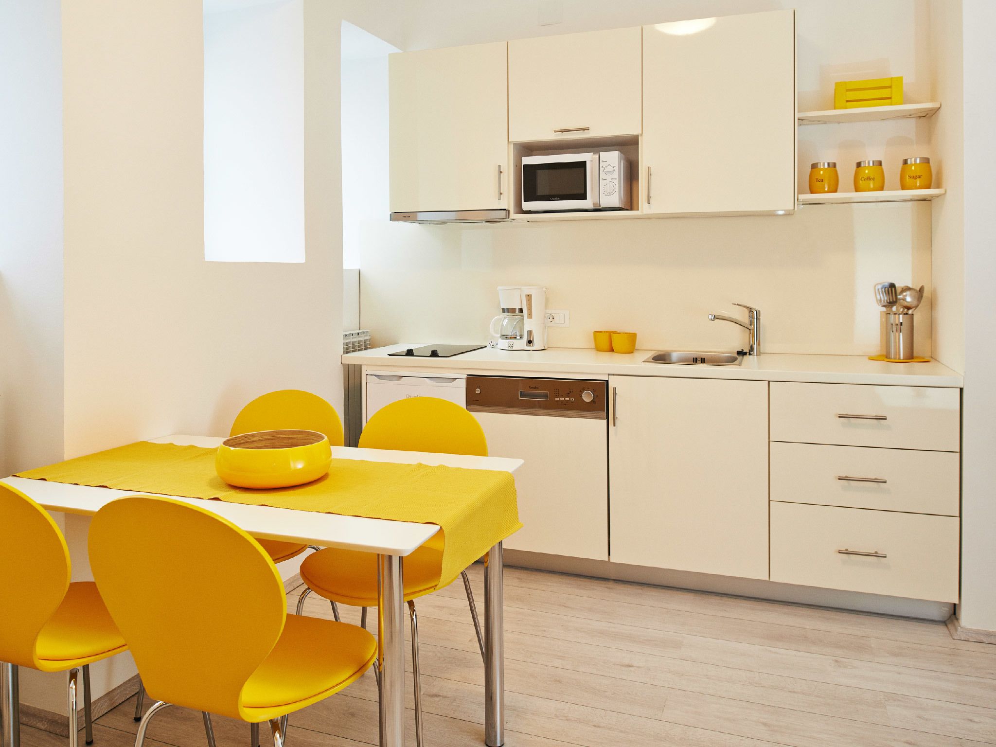 Una cocina compacta con microondas, cafetera y lavamanos. Hay una mesa de comedor con sillas amarillas y un tazón amarillo encima.