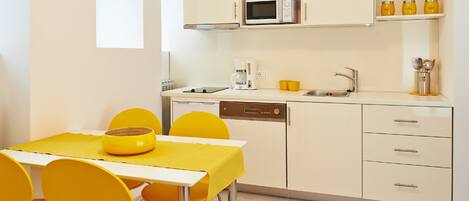 Une cuisine compacte équipée d’un four à micro-ondes, d’une machine à café et d’un évier. On y trouve une table à manger avec des chaises jaunes et un saladier jaune posé dessus.