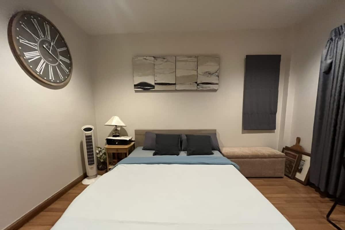 A bedroom with a large bed, a wall clock, a bedside table with a lamp, a small table with a vase, and a window with curtains.