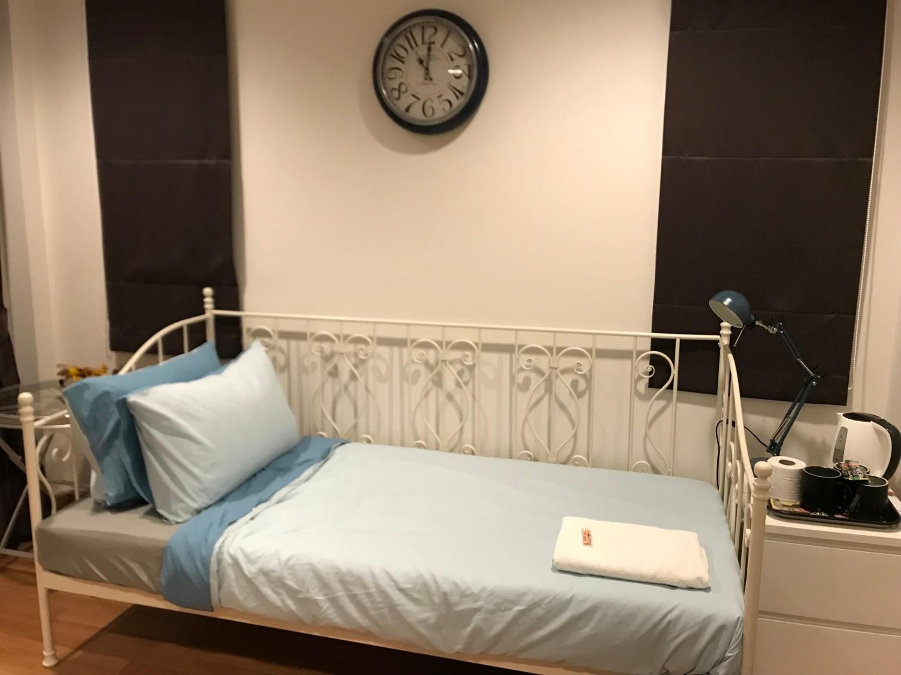 Habitación con una cama individual de estructura metálica blanca, cubres cama azul claro, almohada blanca y un reloj en la pared.