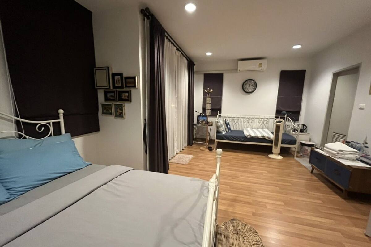 A bedroom with a bed, a bedside table, a small desk with a chair, a window with curtains, and a wall-mounted clock.