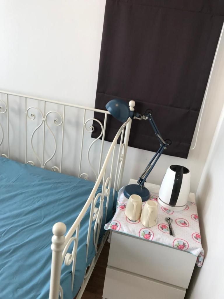 A bed with a blue sheet, a bedside table with a lamp, a cup, and a kettle.