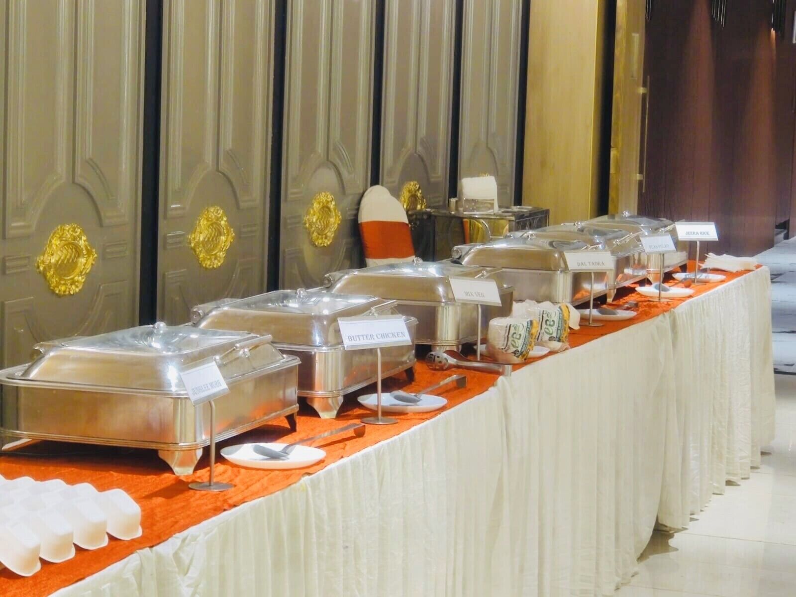 A buffet setup with labeled containers for various food items, including butter, chicken, and salad, on an orange tablecloth.