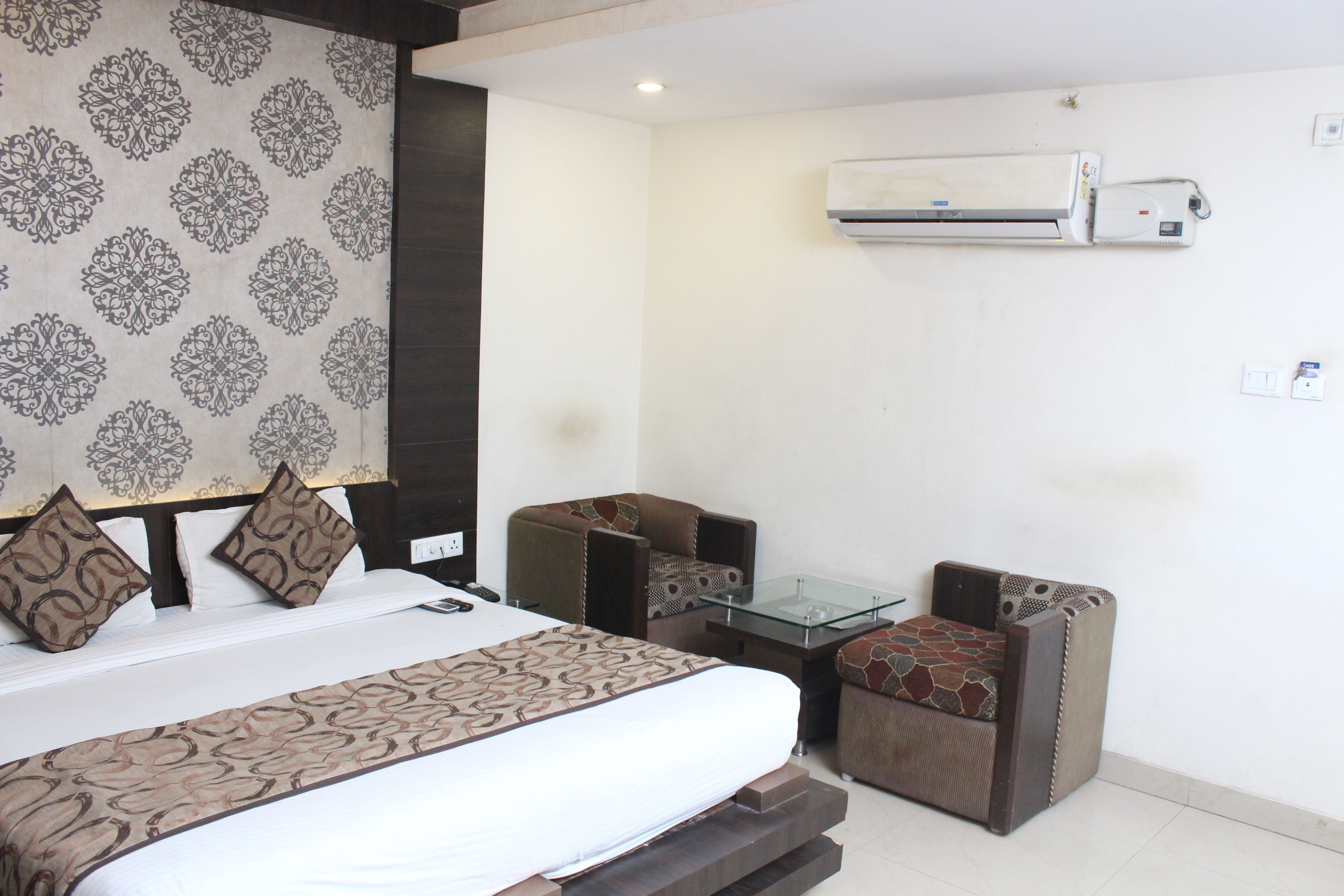 A hotel room with a bed, two armchairs, a glass table, and an air conditioning unit.