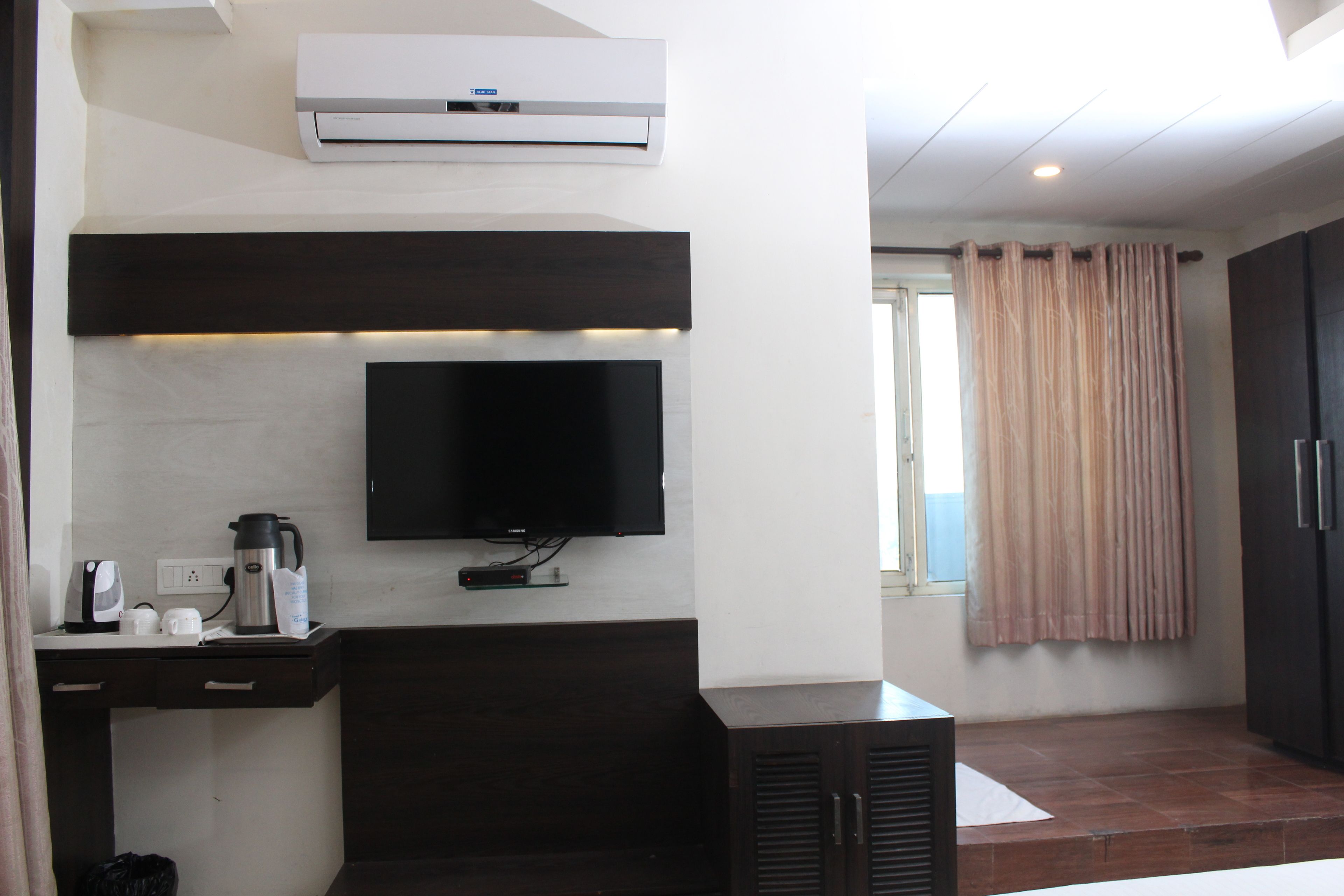 A modern hotel room with a flat-screen TV mounted on the wall, a coffee maker, and a small table with a cup and a remote control.