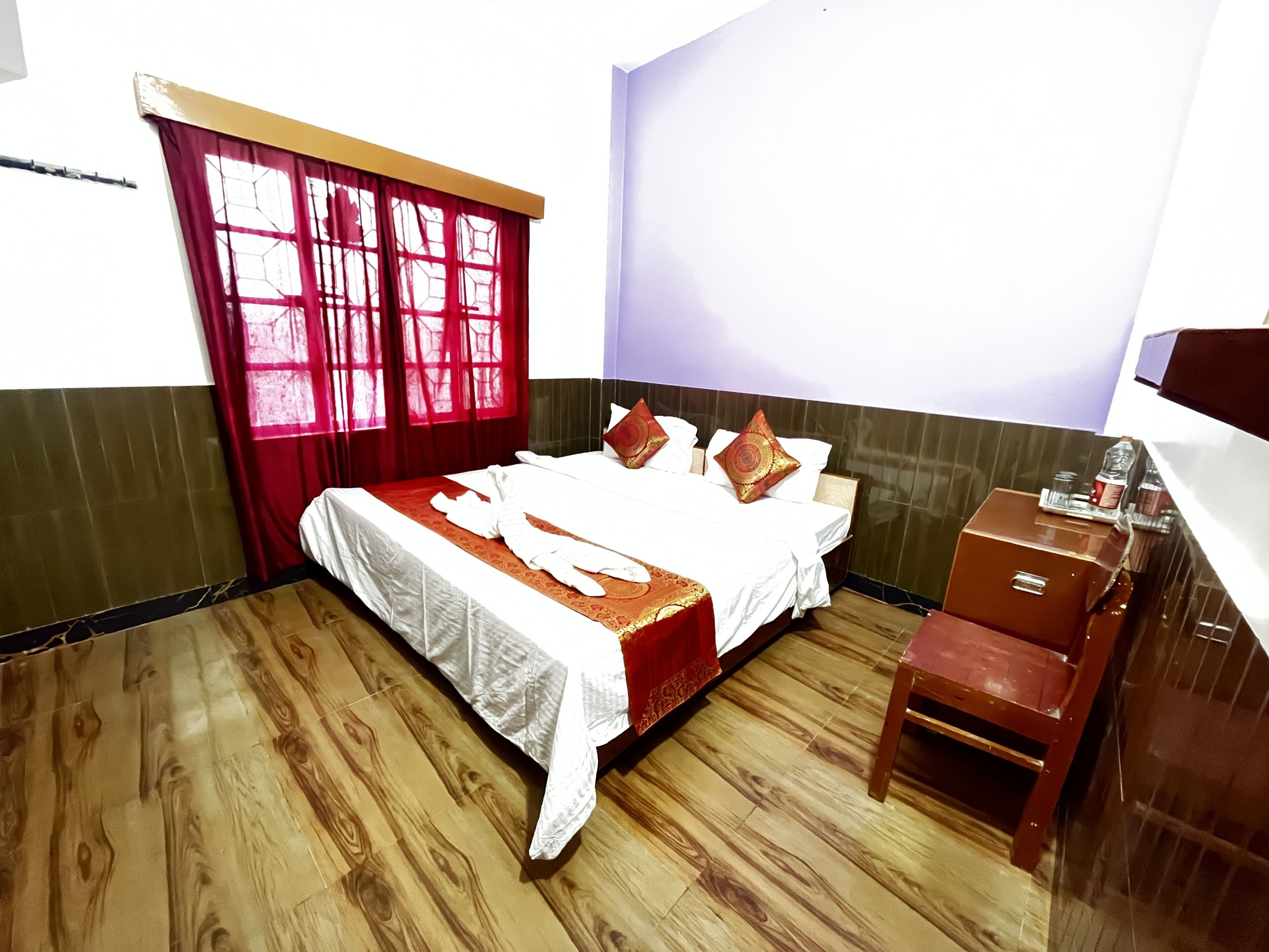 Habitación de hotel con piso de madera, cama con ropa blanca y detalles rojos, mesita de noche de madera y ventana con cortinas rojas.
