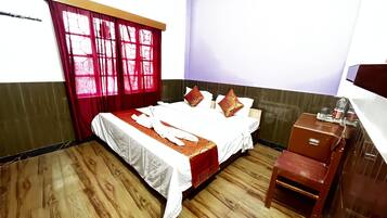 Habitación de hotel con piso de madera, cama con ropa blanca y detalles rojos, mesita de noche de madera y ventana con cortinas rojas.