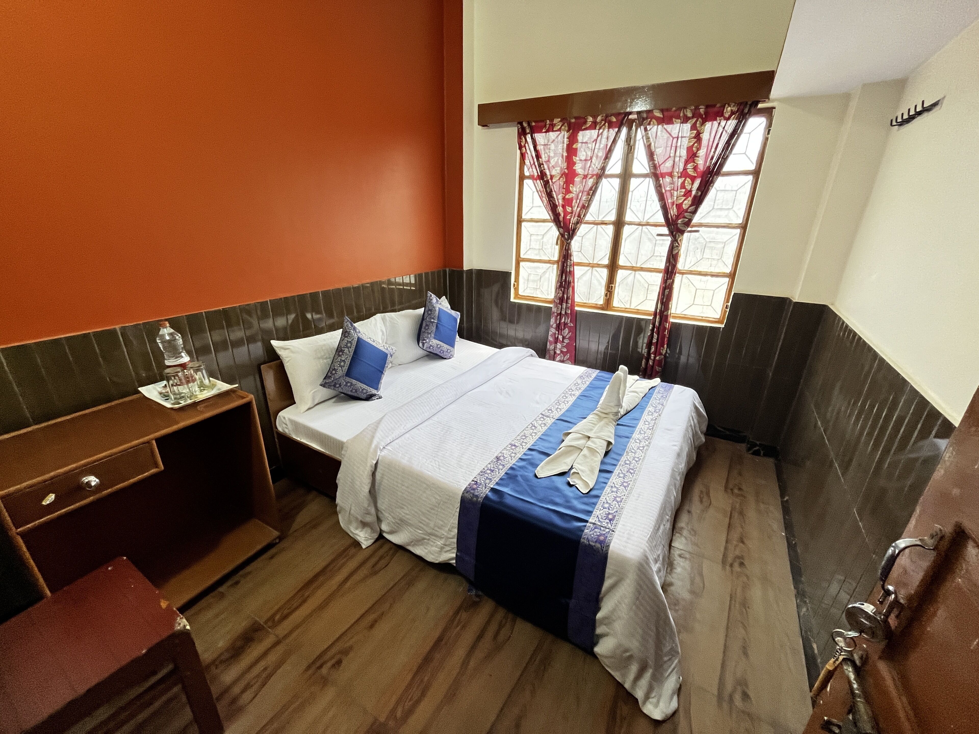 A hotel room with a bed, a wooden desk, a window with curtains, and a wall with a decorative pattern.