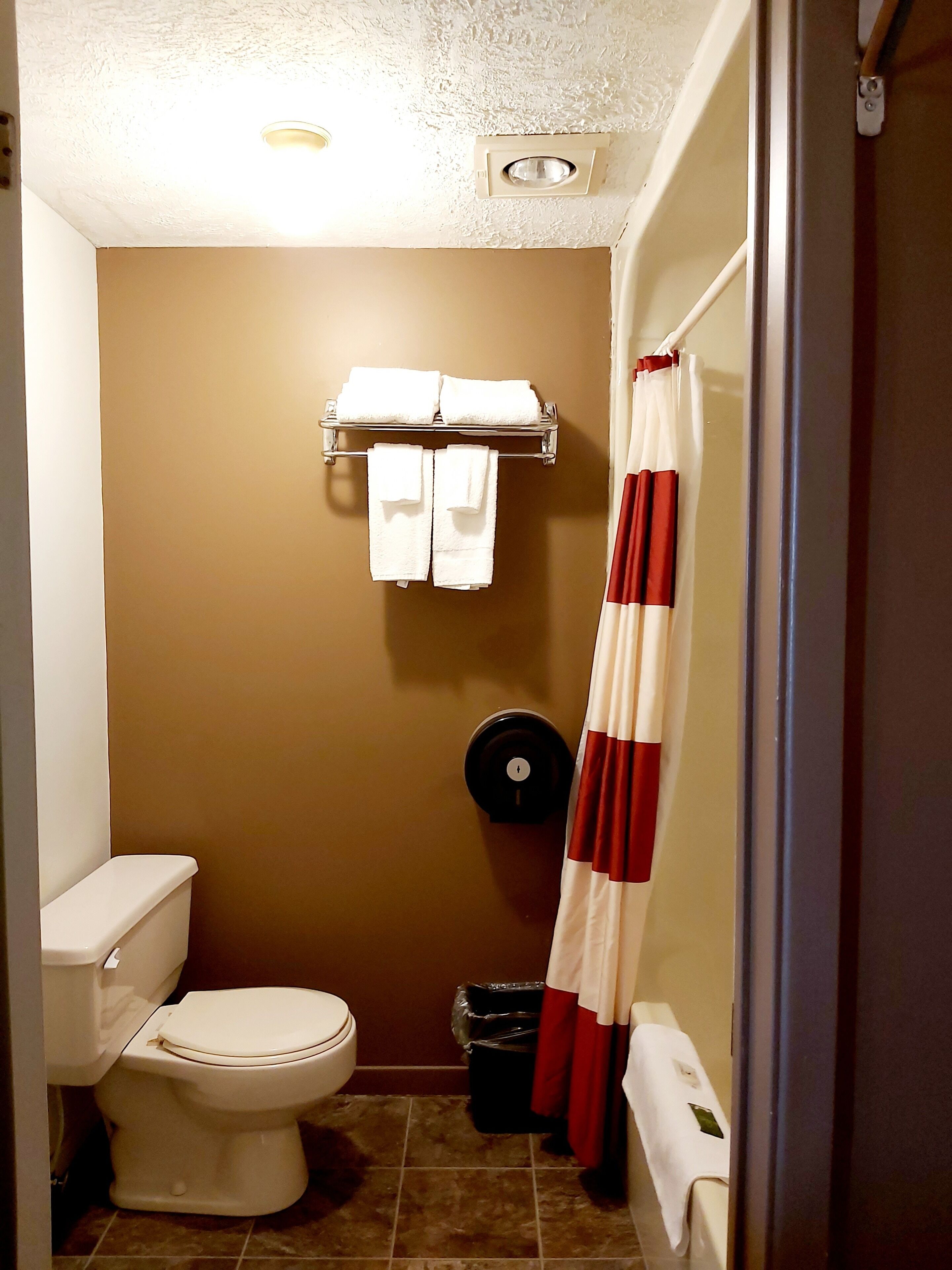 A small bathroom with a toilet, a towel rack with folded towels, a trash bin, and a shower curtain.