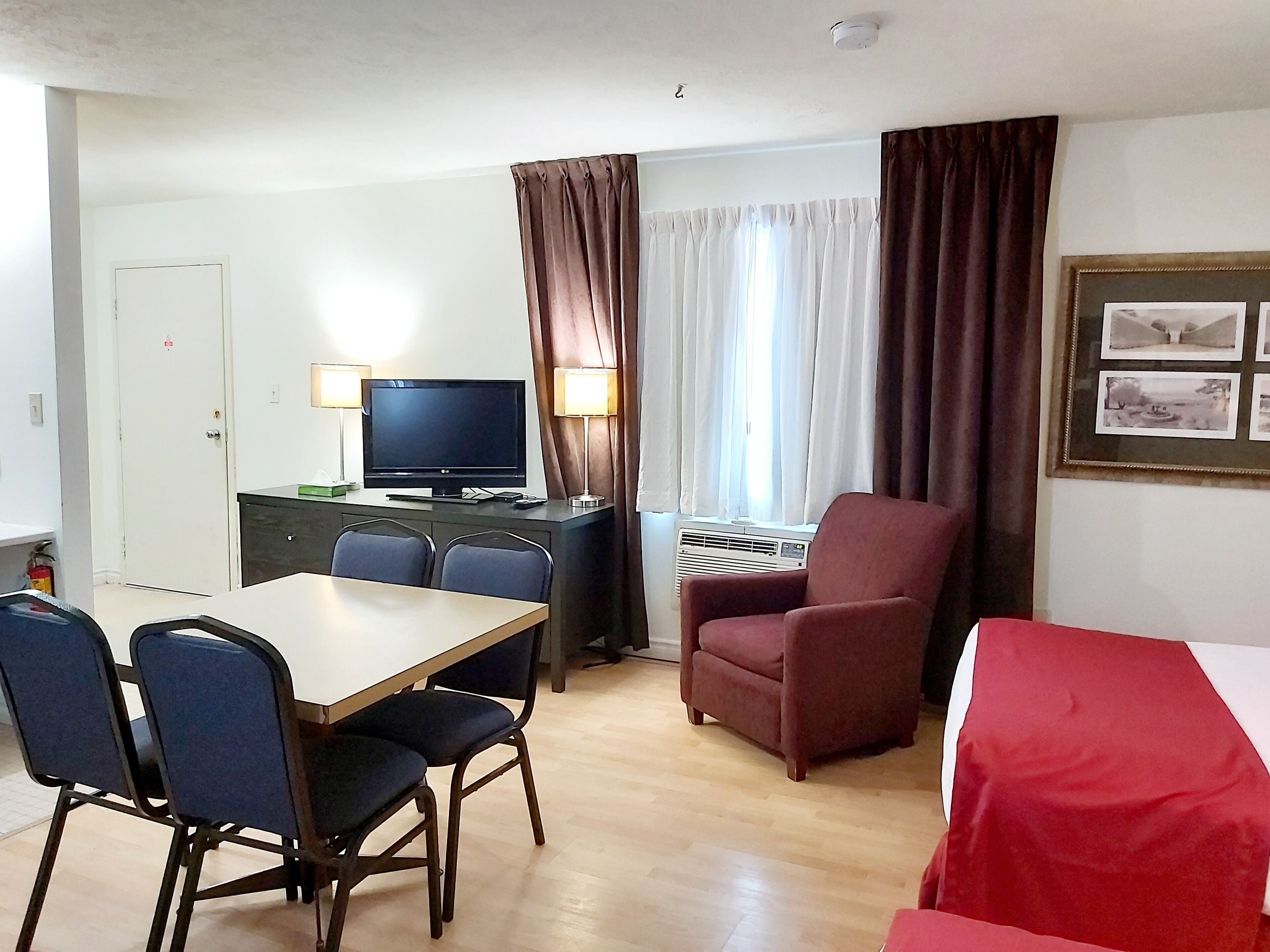 Une chambre d’hôtel avec une table à manger en bois, deux chaises bleues, un fauteuil rouge, une télévision, un bureau avec une lampe et un tableau encadré au mur.