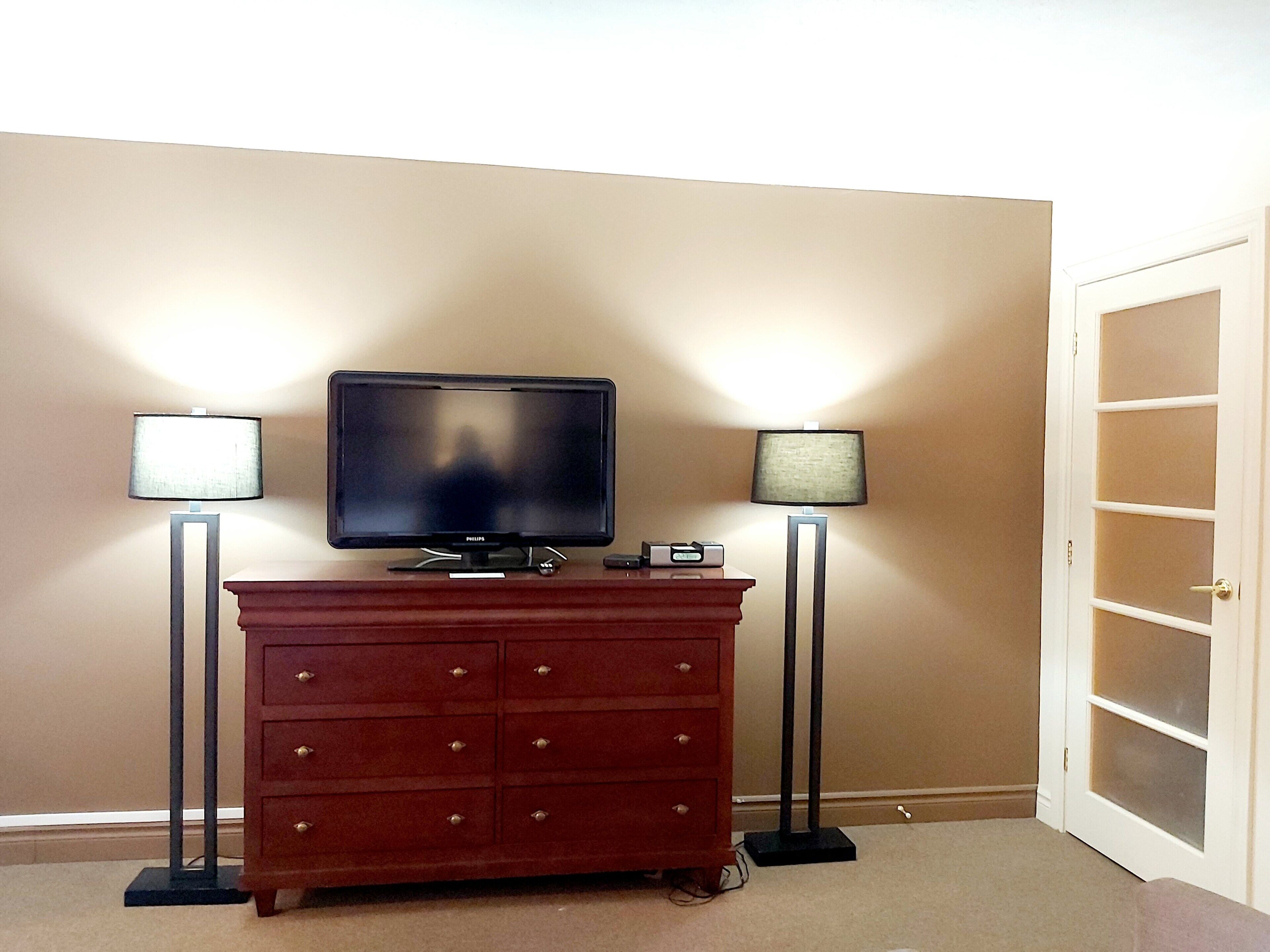 A room with a television on a wooden dresser, two floor lamps, and a door with frosted glass panels.