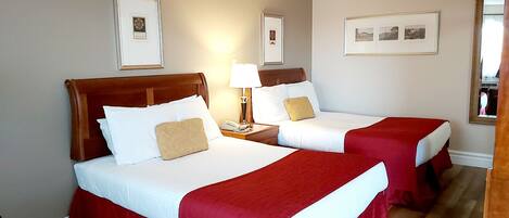 Une chambre d’hôtel avec deux lits, chacun recouvert d’une couverture rouge et doté de draps blancs. Des tableaux encadrés ornent les murs, et un miroir se trouve sur le côté droit.