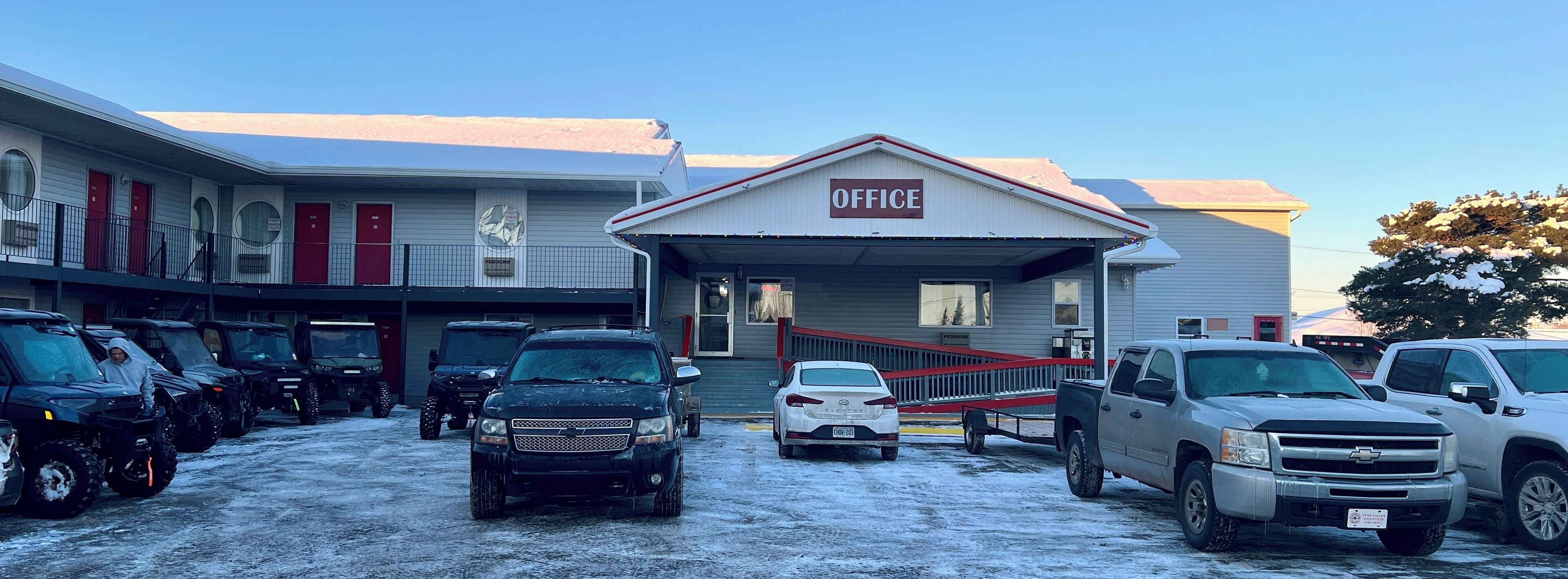A building with an 'OFFICE' sign, a covered entrance, and a parking lot with various vehicles.