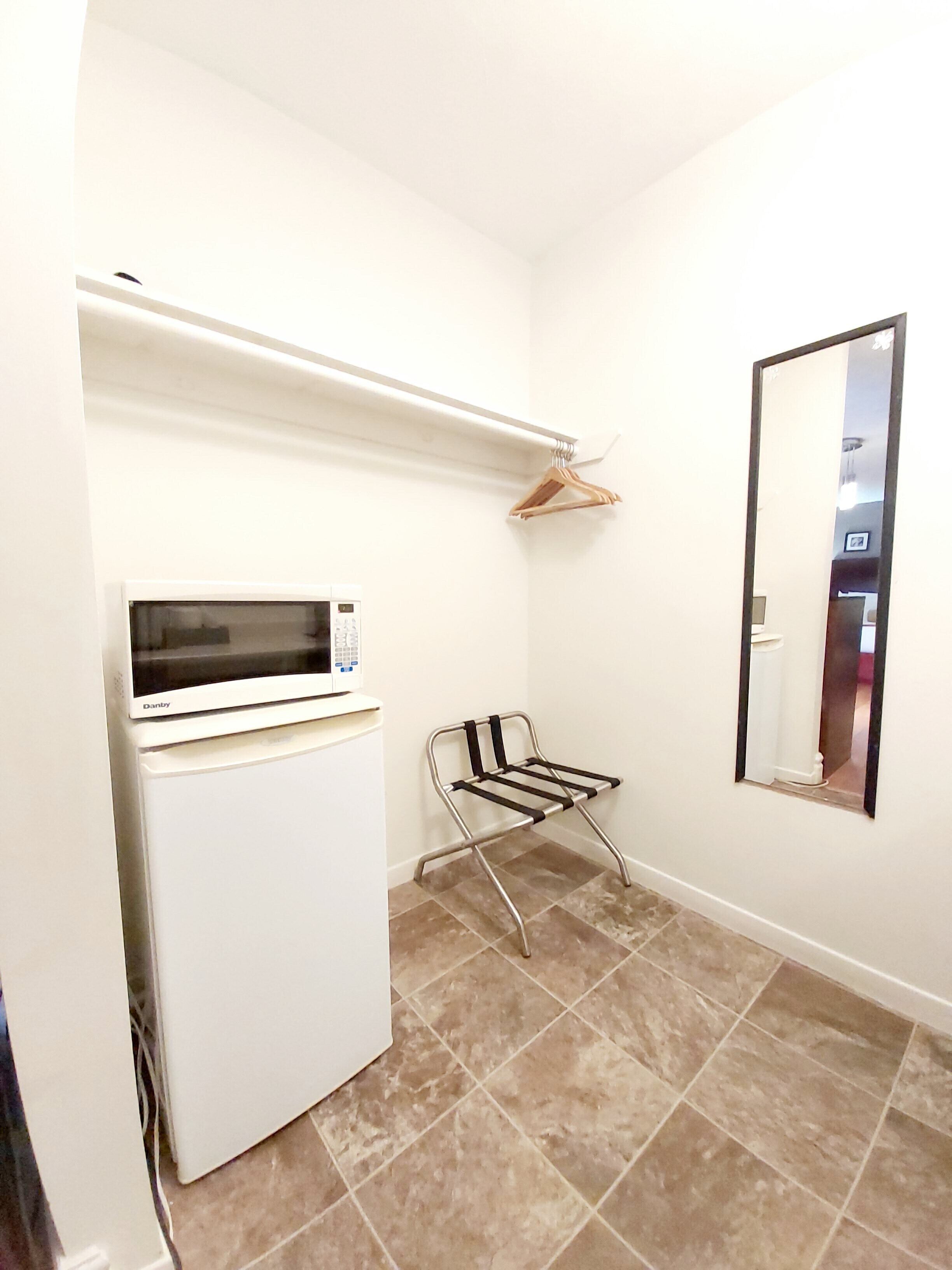 A small, clean room with a white refrigerator, a microwave, a metal drying rack, and a mirror on the wall.