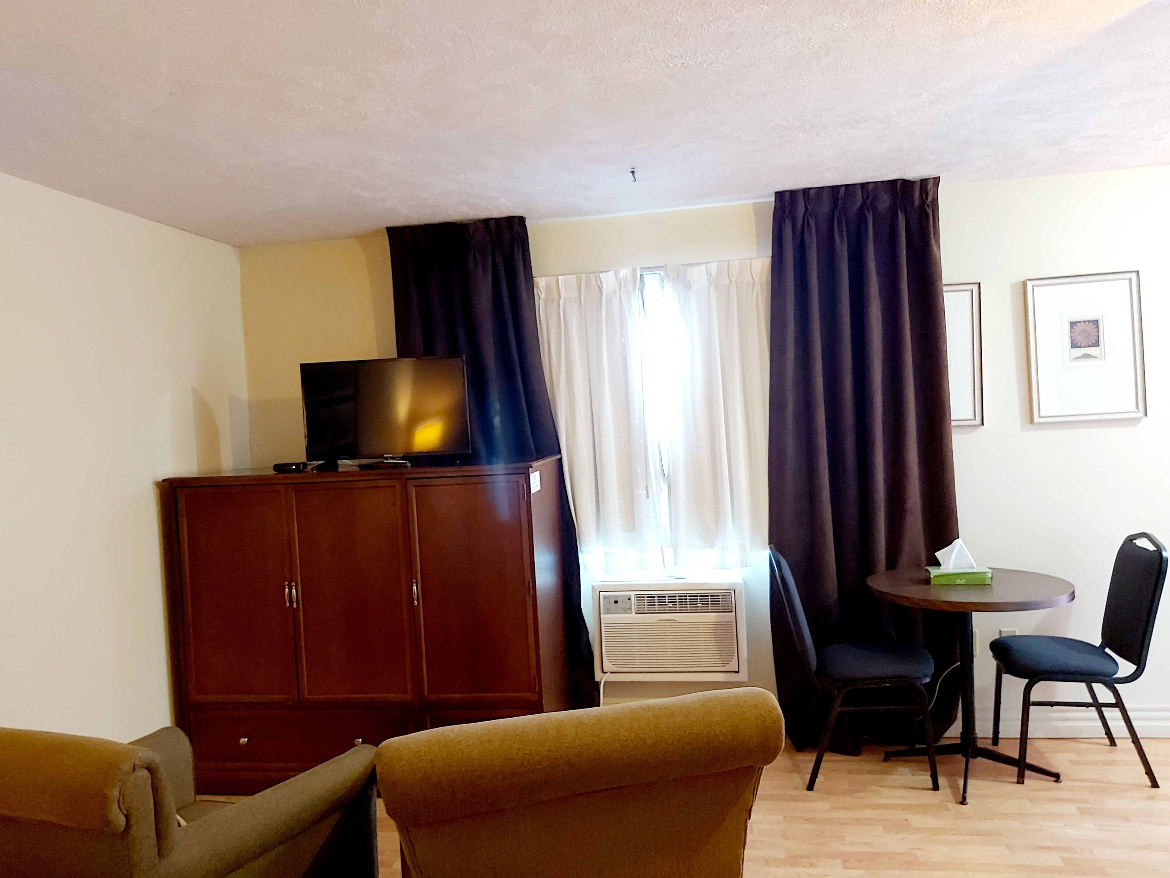 A room with a TV on a wooden cabinet, a table with chairs, a window with curtains, and an air conditioning unit.