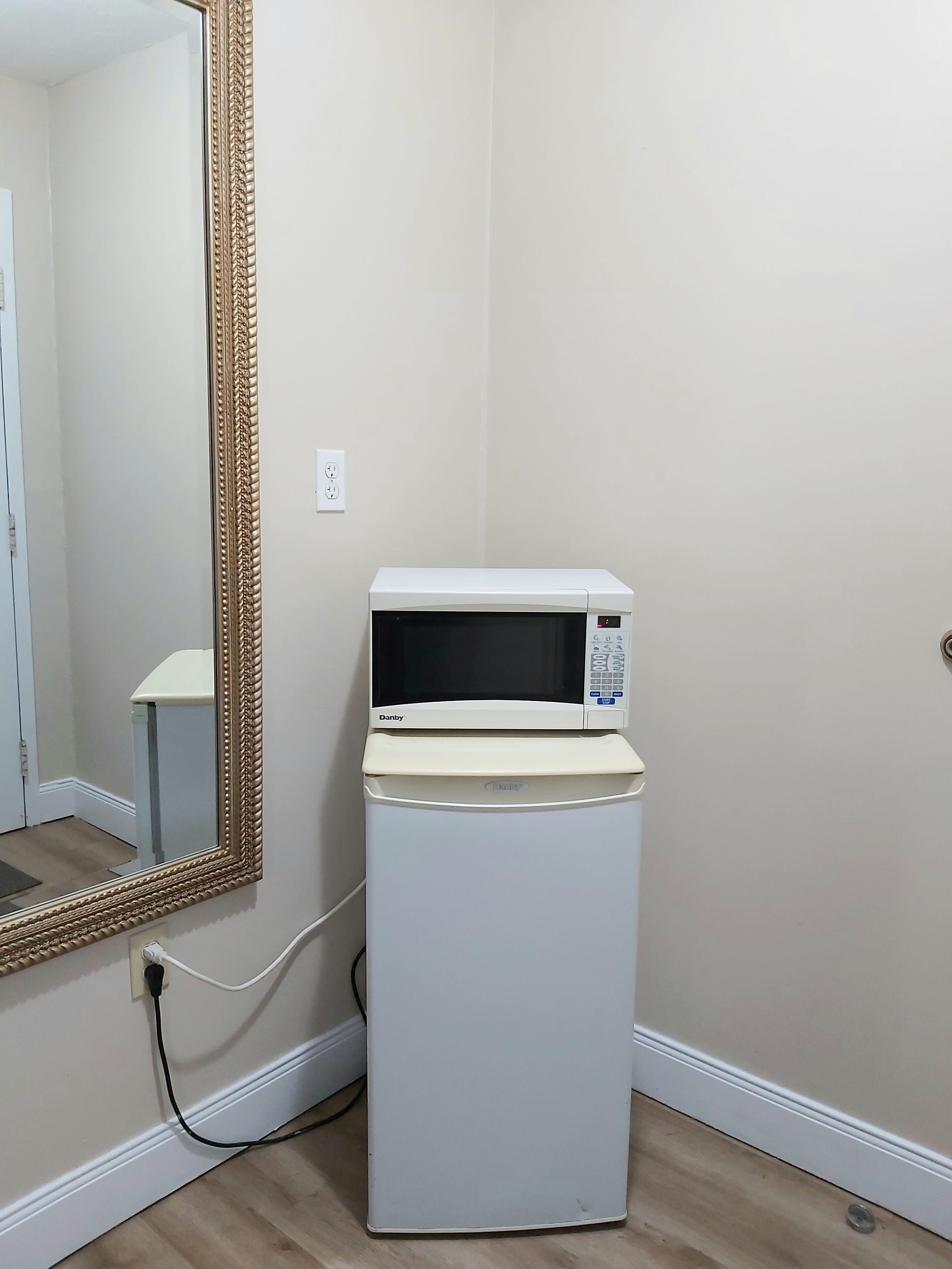 A compact white microwave on a white stand in a corner with a large ornate mirror on the wall.